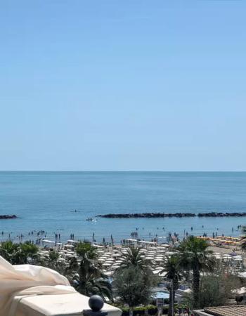 Spiaggia affollata con ombrelloni, palme e mare calmo sotto un cielo sereno.