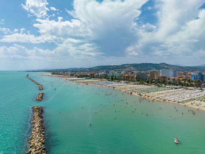 Spiaggia affollata con ombrelloni, mare turchese e colline sullo sfondo sotto un cielo nuvoloso.